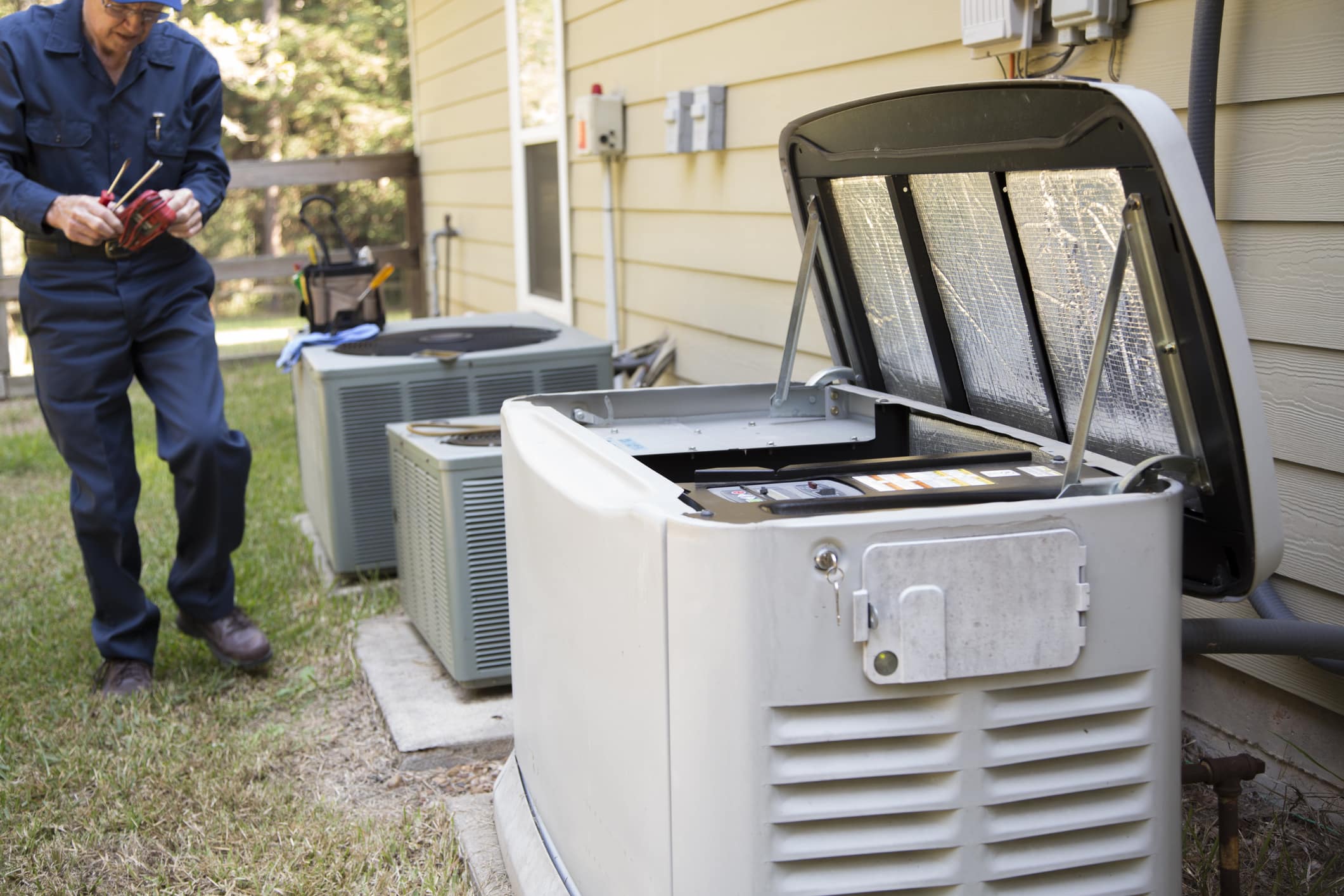An electrician approaches a home generator against the side of a house to inspect their electrical work.