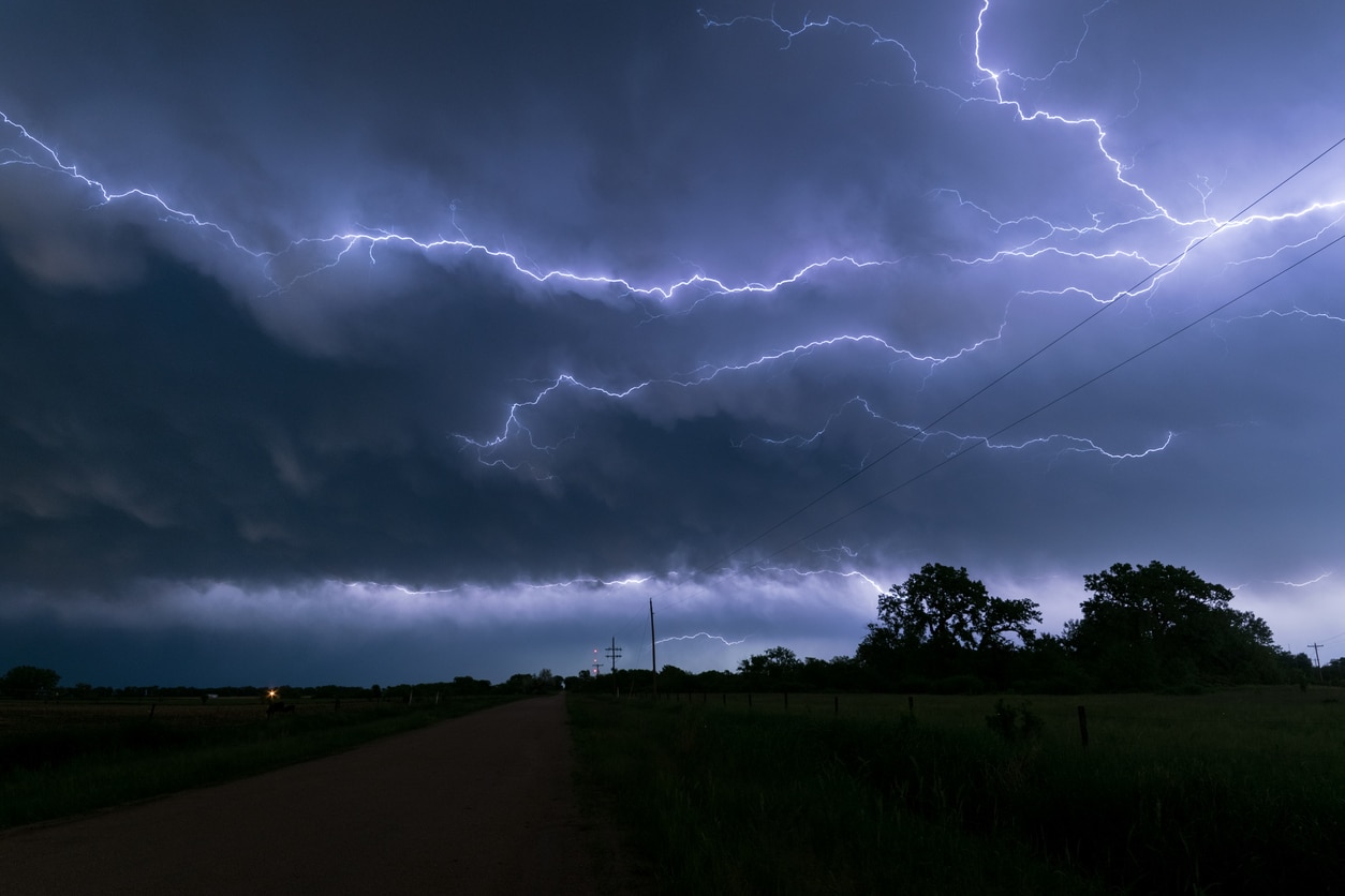 A dark sky with a strike of lightning over a field with trees and powerlines.