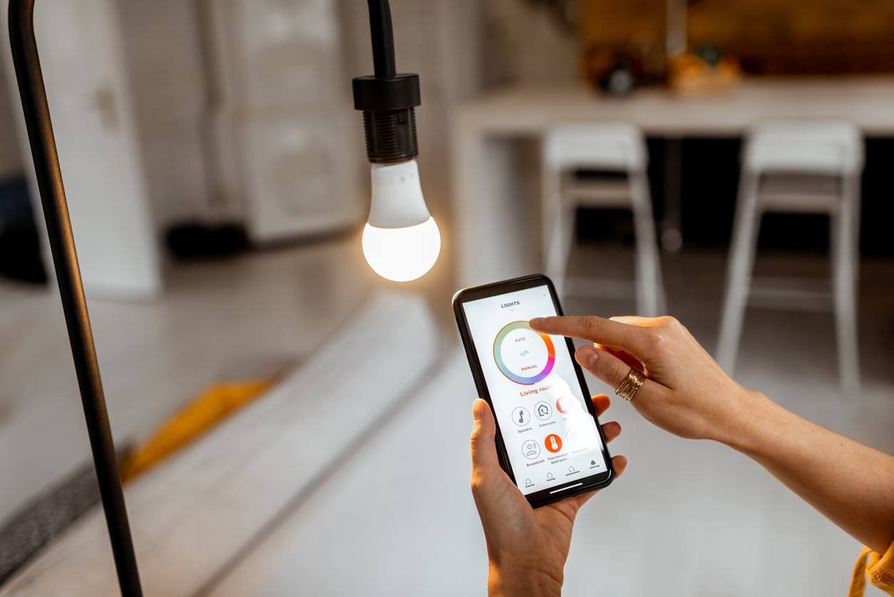 Person adjusting smart home lighting on smartphone app next to a glowing hanging light bulb in a modern room