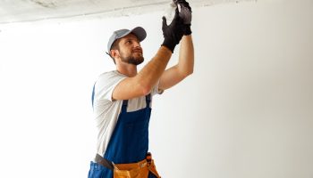 A licensed electrician wearing gloves while installing a ceiling light bulb in a room with exposed wiring.