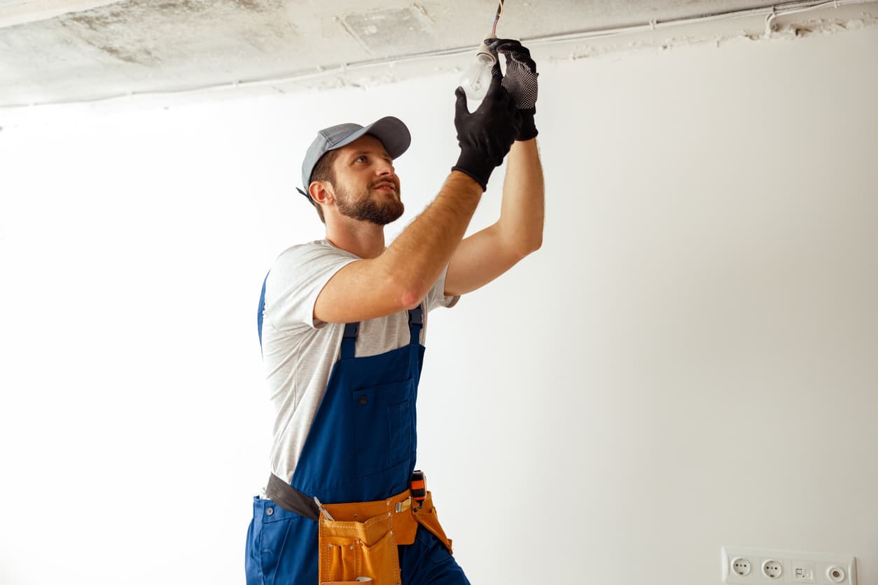 A licensed electrician wearing gloves while installing a ceiling light bulb in a room with exposed wiring.