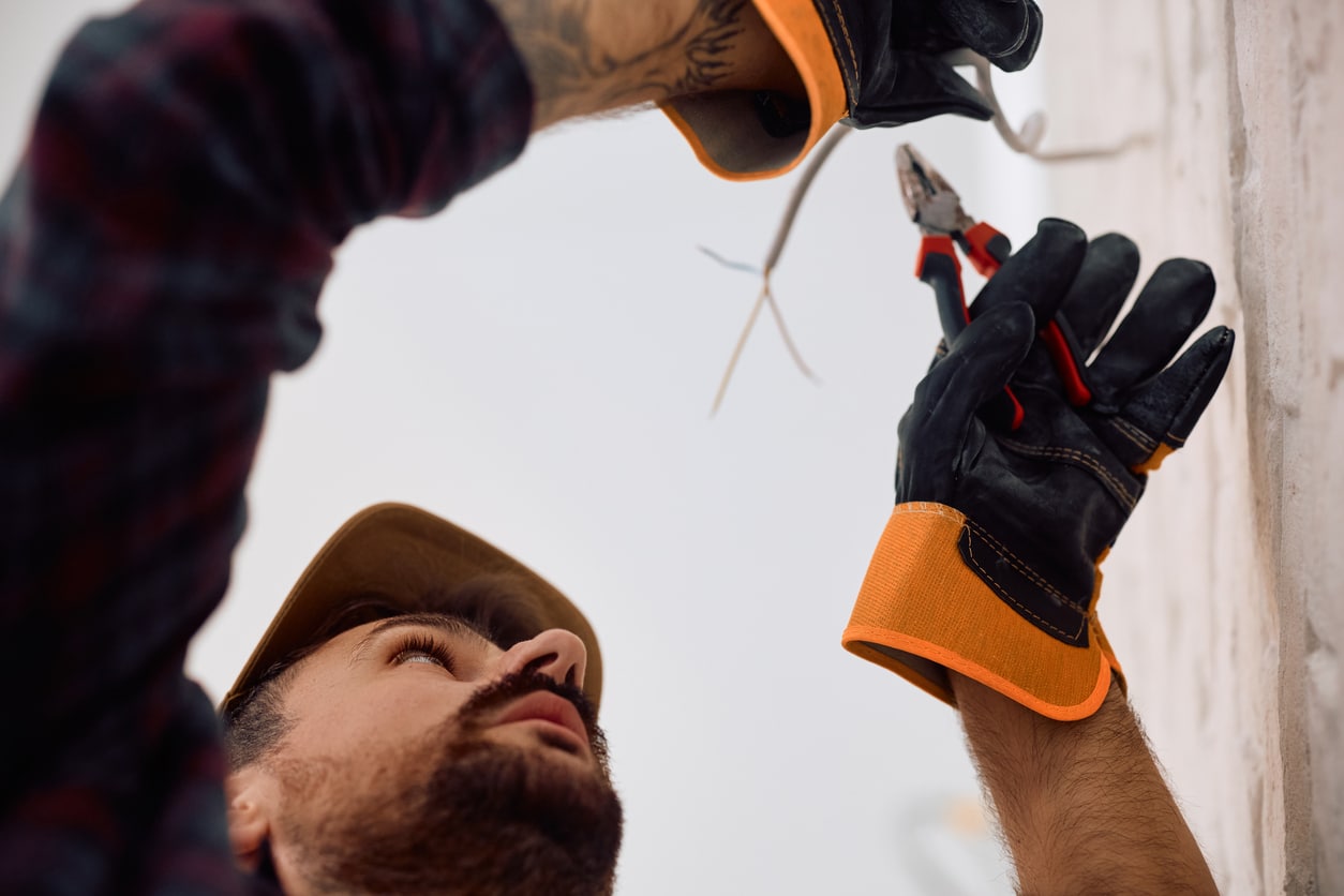 A professional electrician in gloves using pliers to cut or strip exposed wires on a wall during electrical repair.
