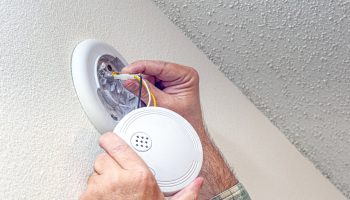 Hands wiring a hardwired smoke detector into its ceiling mount, connecting the wire harness before securing the unit.